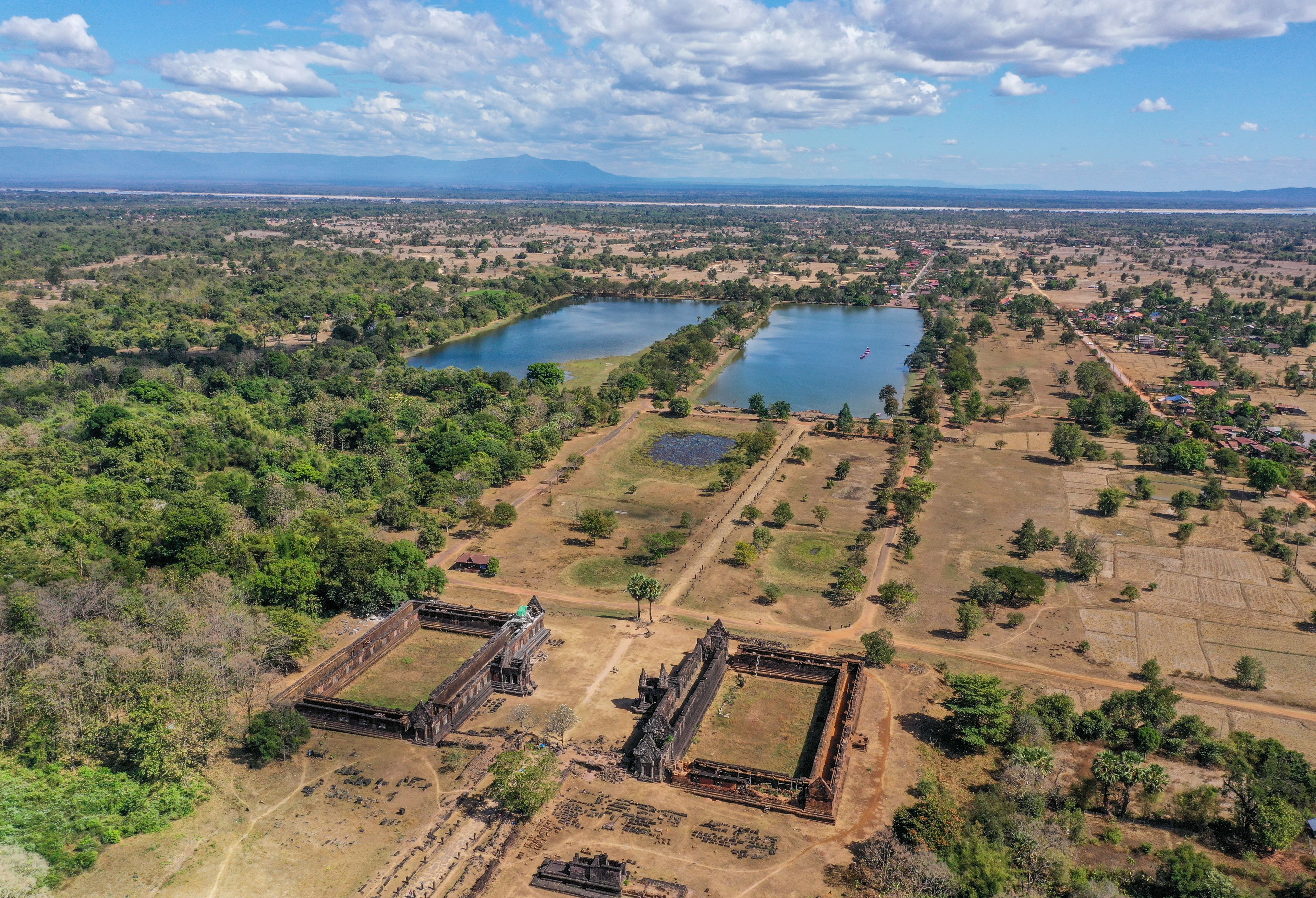 An aerial view of a historical site with large open courtyards, a lake, and surrounding greenery.