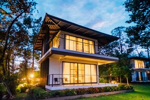 Una casa moderna de dos pisos con ventanas grandes y un balcón, rodeada de follaje.