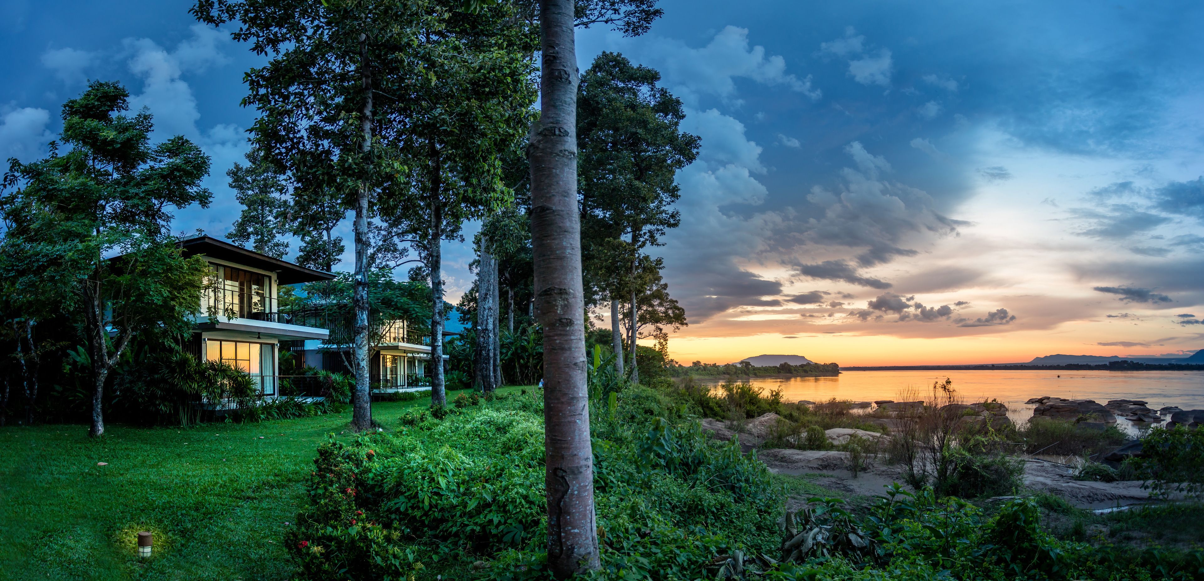 A modern house with a large glass facade, surrounded by lush greenery and a view of a calm lake at sunset.
