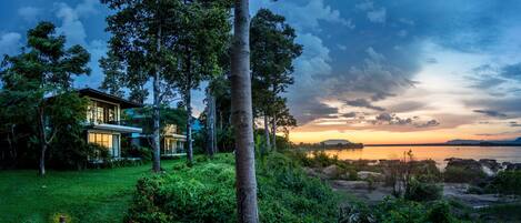 A modern house with a large glass facade, surrounded by lush greenery and a view of a calm lake at sunset.