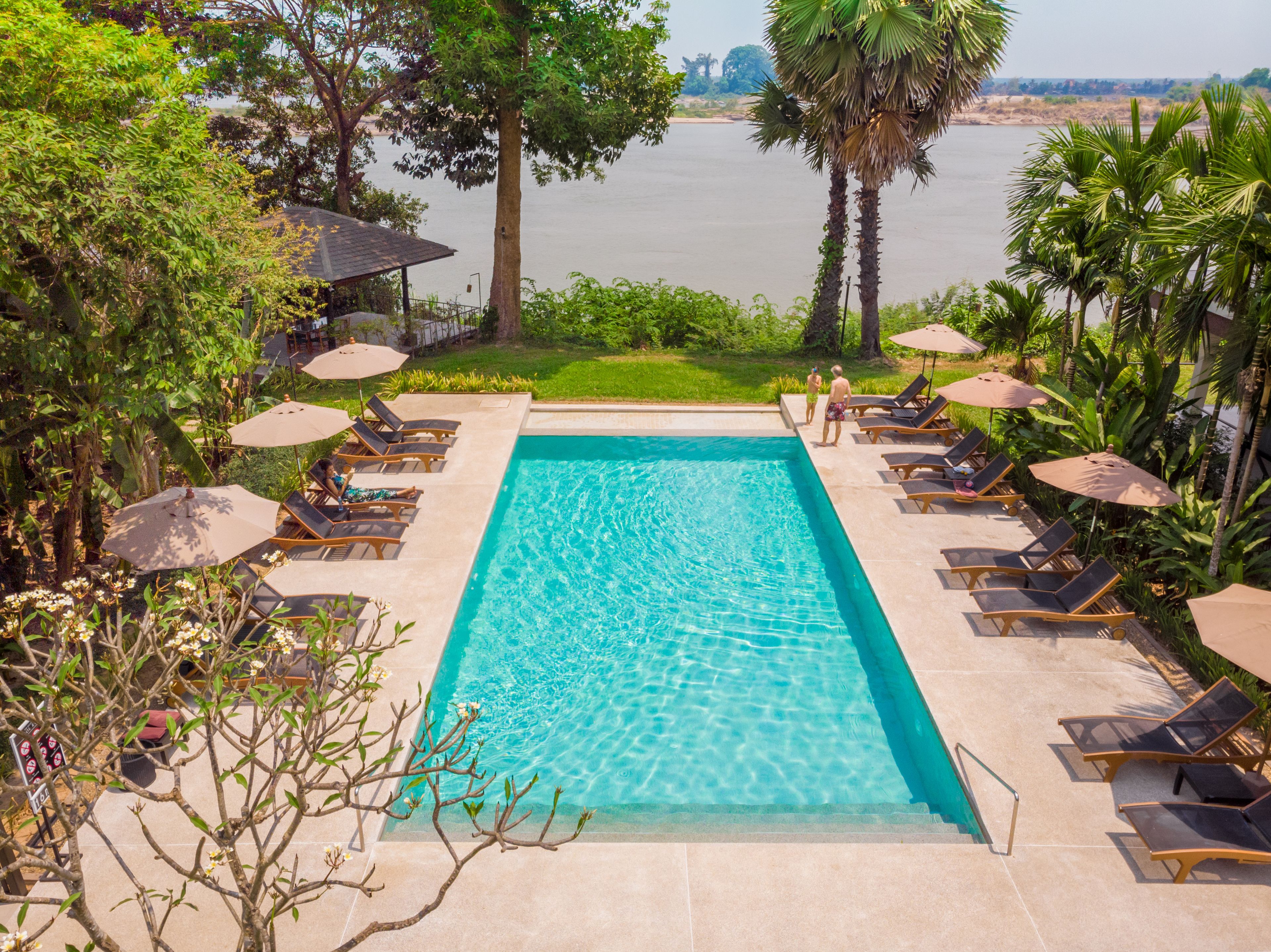A rectangular pool with clear blue water, surrounded by lounge chairs and umbrellas, with a view of a river and greenery in the background.