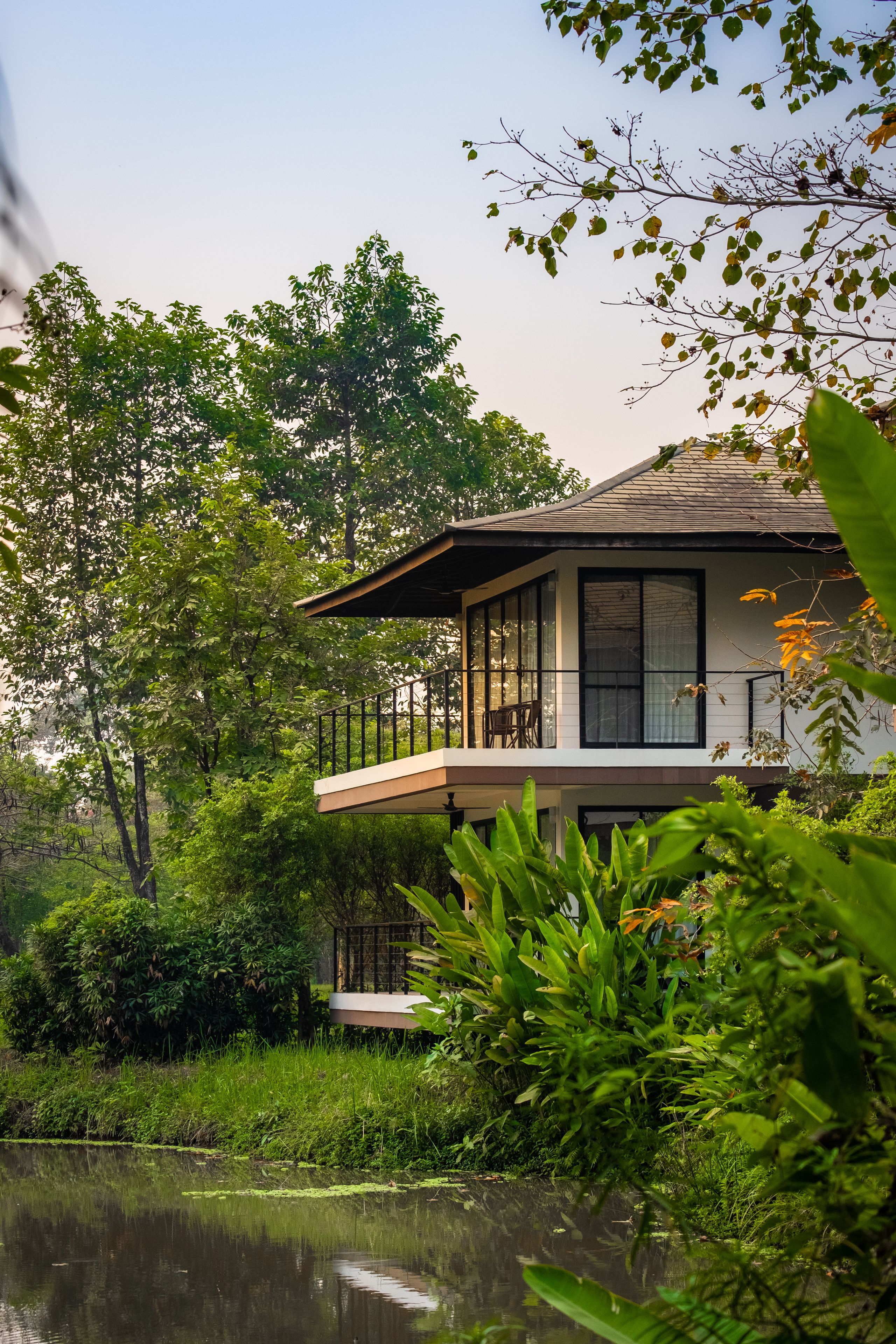 A house with a balcony overlooking a pond, surrounded by lush greenery.