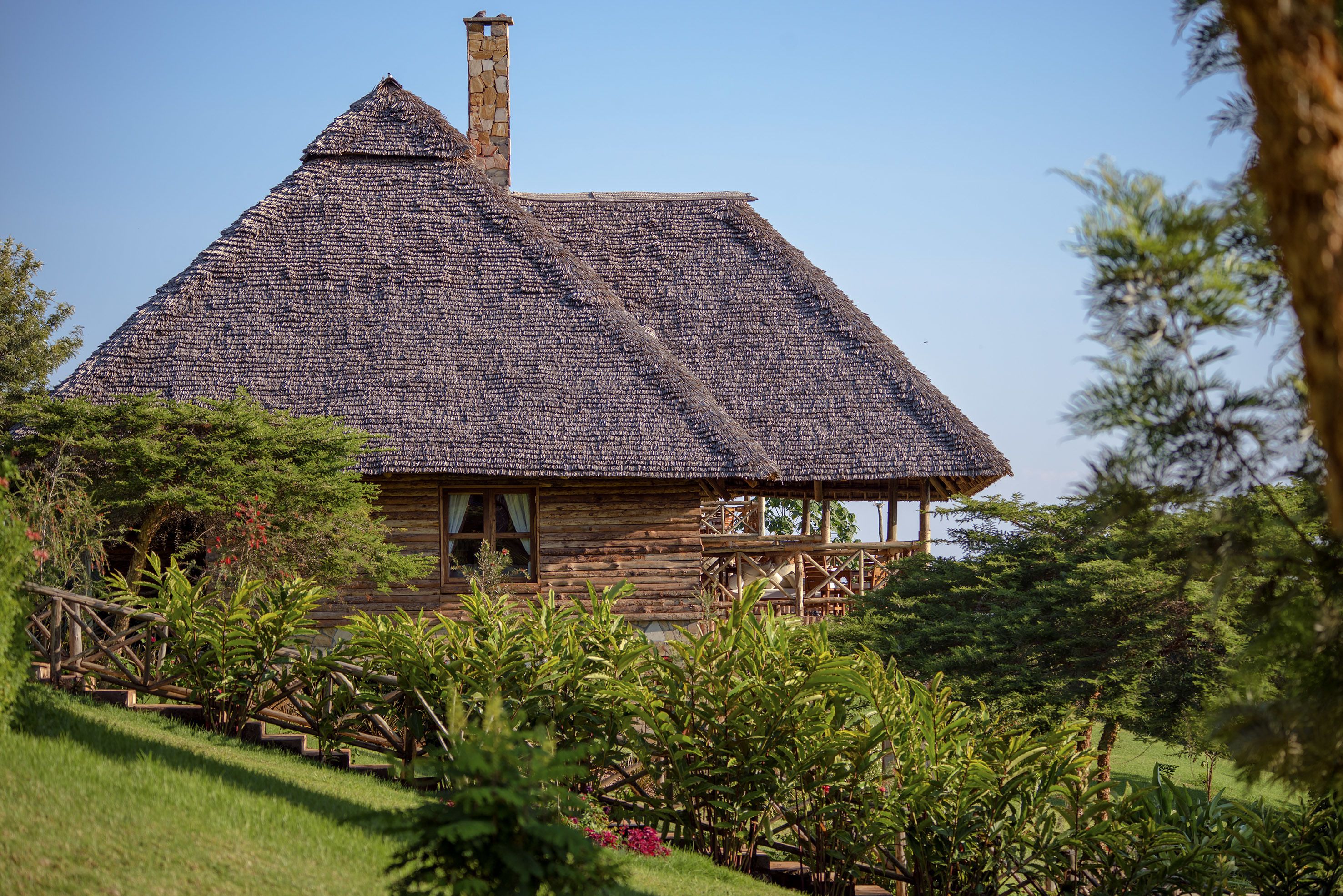 Une cabane rustique en rondins de bois, dotée d’un toit de chaume à forte pente, entourée d’une végétation luxuriante et agrémentée d’une terrasse en bois.