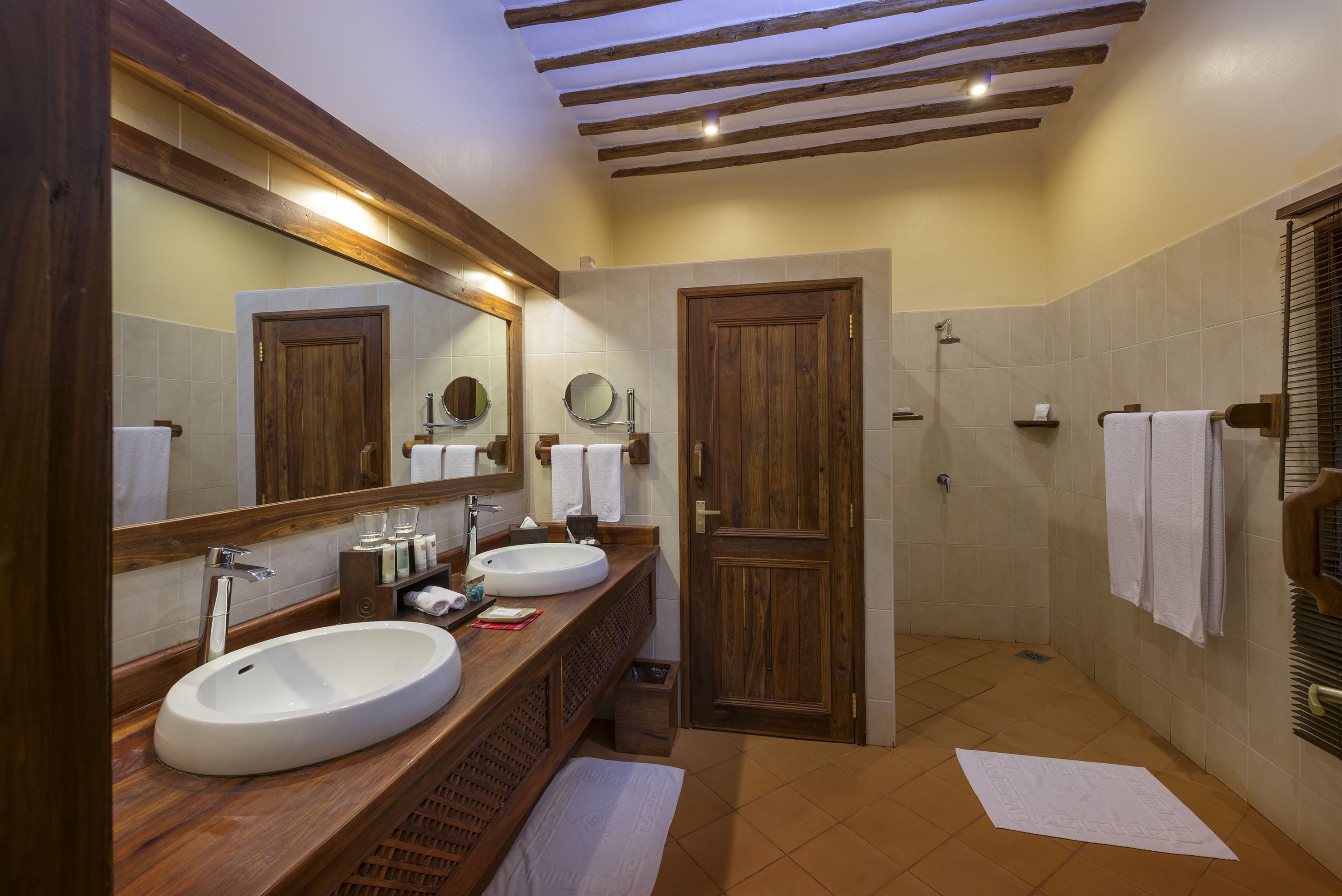 A bathroom with two sinks, a large mirror, wooden beams on the ceiling, and a shower area.