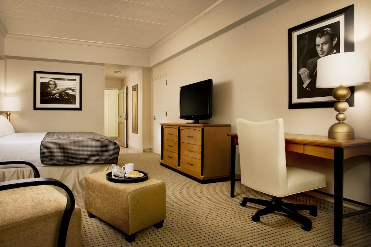 A hotel room with a bed, a desk, a chair, a television, a dresser, and a small table with a tray.
