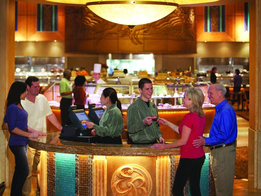 A hotel lobby with a large, ornate emblem above a reception desk, a chandelier, and a view of the dining area in the background.