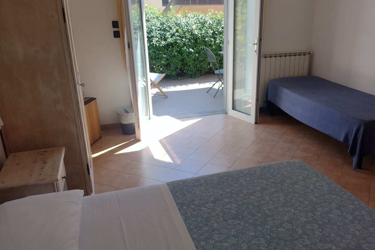 A small, clean room with a bed, a chair, and a table. There is a window with a view of a garden and a door leading outside.