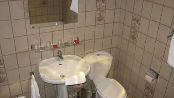 A bathroom with a sink, toilet, and mirror. There are toiletries on a shelf, and a trash can is visible.