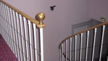 Una escalera con balaustres blancos y barandilla de madera, que lleva a un rellano con un pequeño objeto negro en la pared.