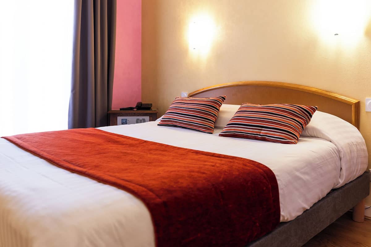 A neatly made bed with a red blanket and two striped pillows, a bedside table with a phone and a lamp, and a wall-mounted light fixture.