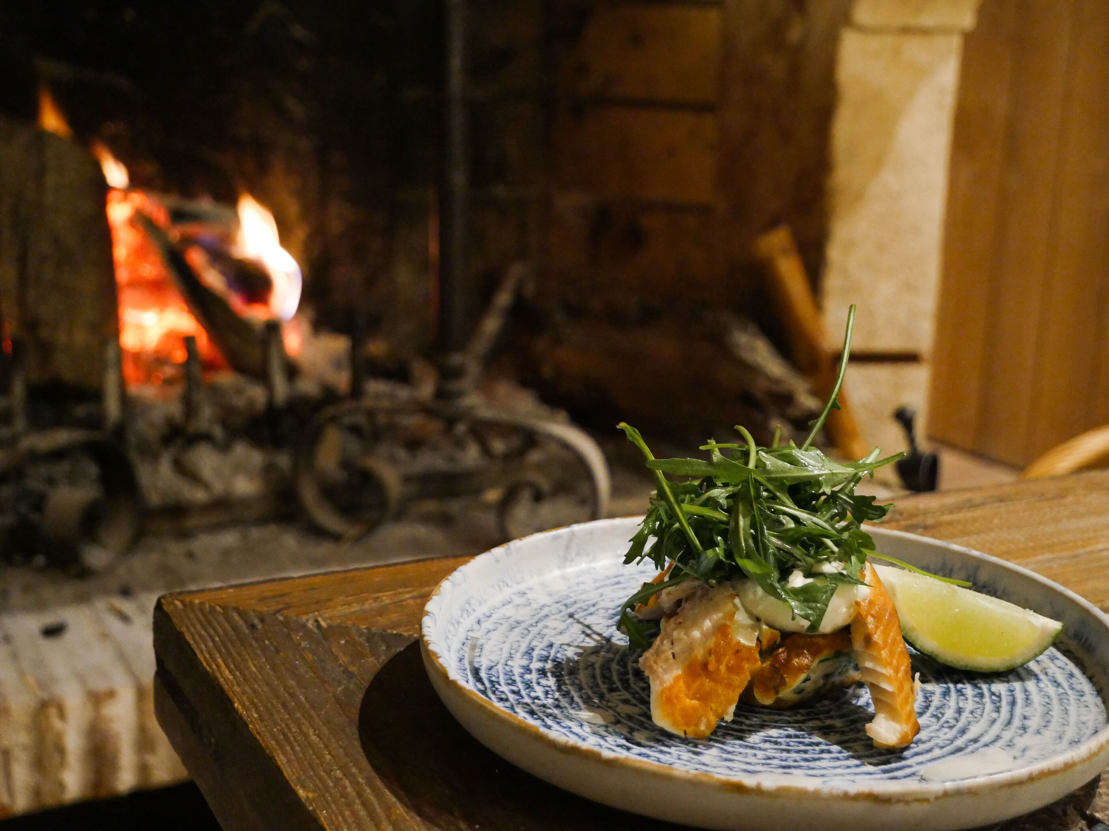 A plate of food with a slice of lime on a patterned plate, set on a wooden table with a fireplace in the background.