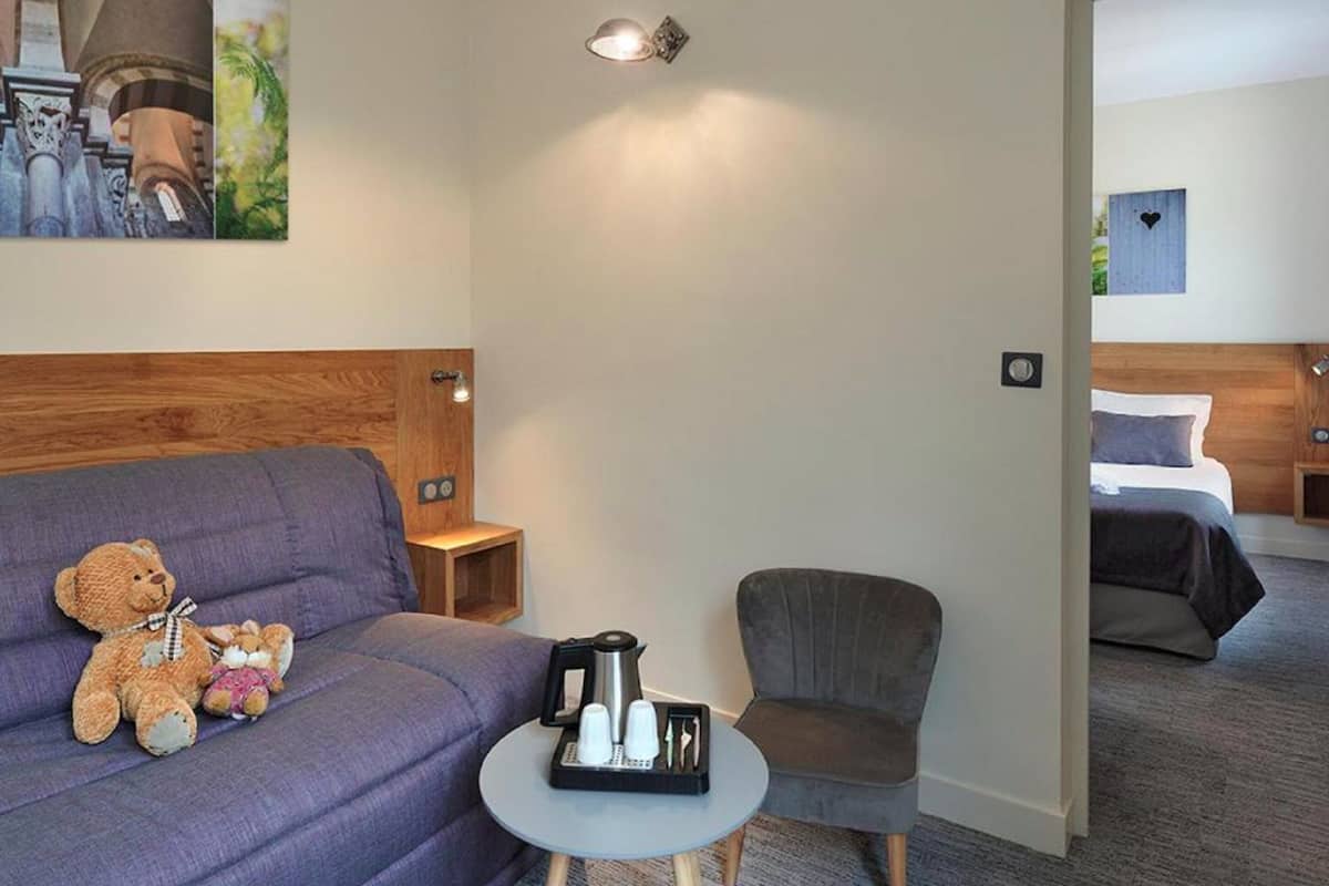 Une chambre d’hôtel avec un canapé violet, un petit fauteuil, une table basse sur laquelle se trouve un plateau avec des tasses et une bouilloire, et un lit à l’arrière-plan.
