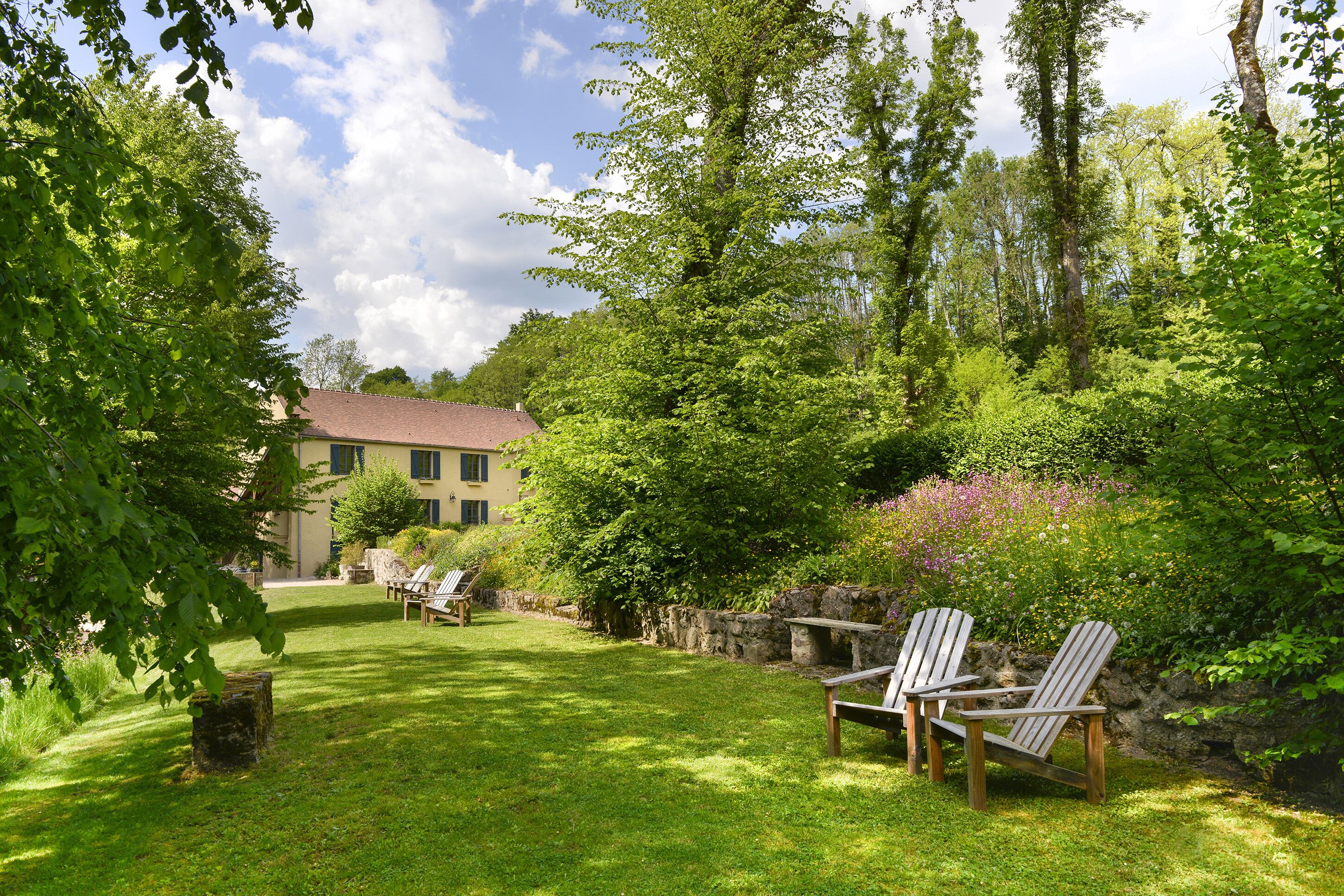 Un jardin bien entretenu, avec un mur en pierre, deux fauteuils Adirondack et, au fond, un bâtiment doté d’un toit rouge.