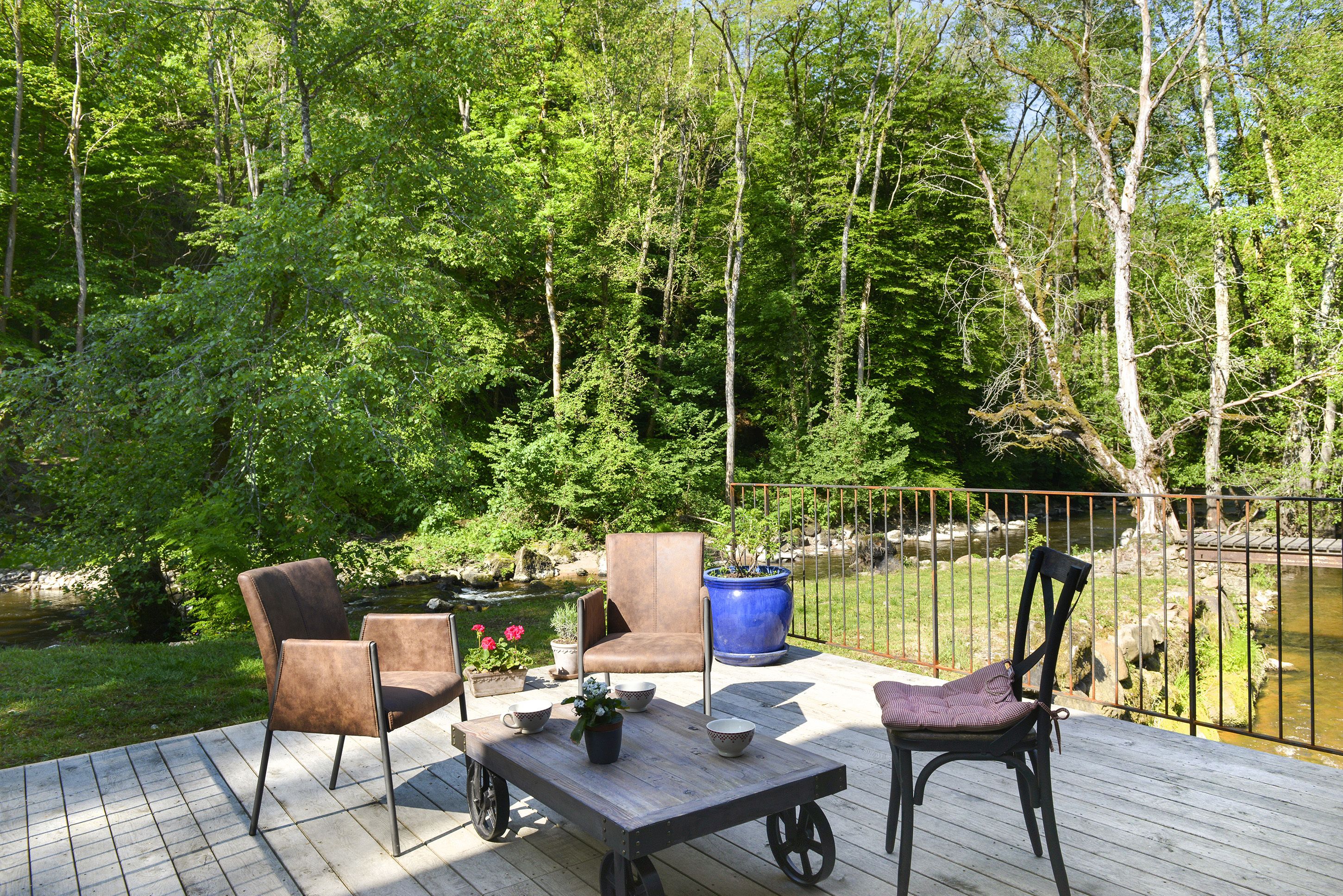 A wooden deck with outdoor furniture, including chairs and a table, overlooking a pond and surrounded by trees.