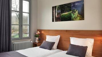 A hotel room with two beds, a window with curtains, a radiator, and a wall-mounted artwork.