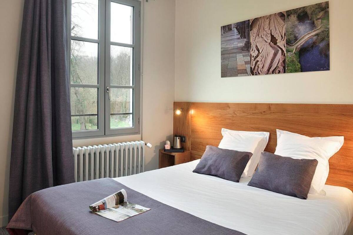 Une chambre d’hôtel avec un grand lit, une fenêtre avec des rideaux, un radiateur, une table de chevet avec une lampe et un tableau au mur.
