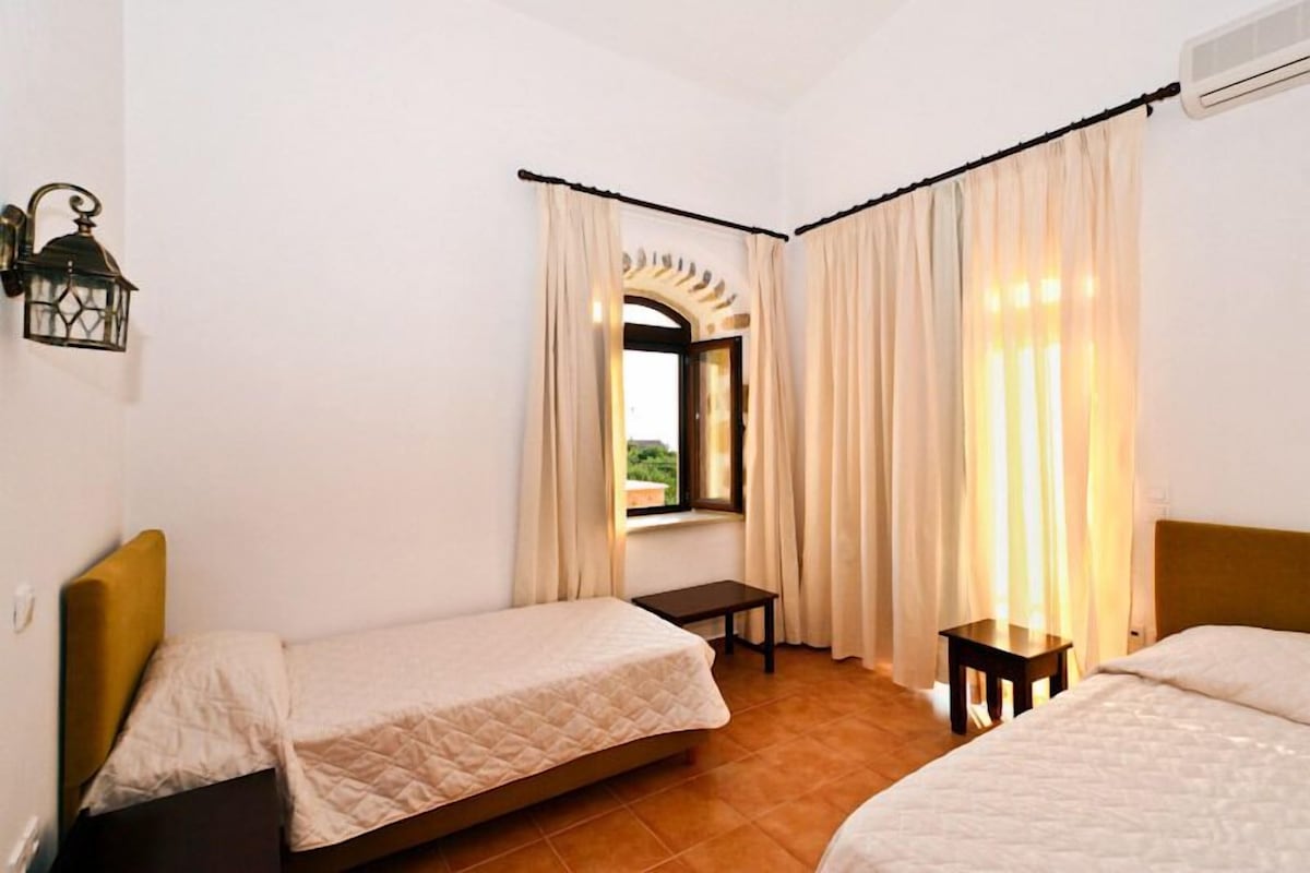 A room with two beds, a window with curtains, a small table, and a wall-mounted light fixture.