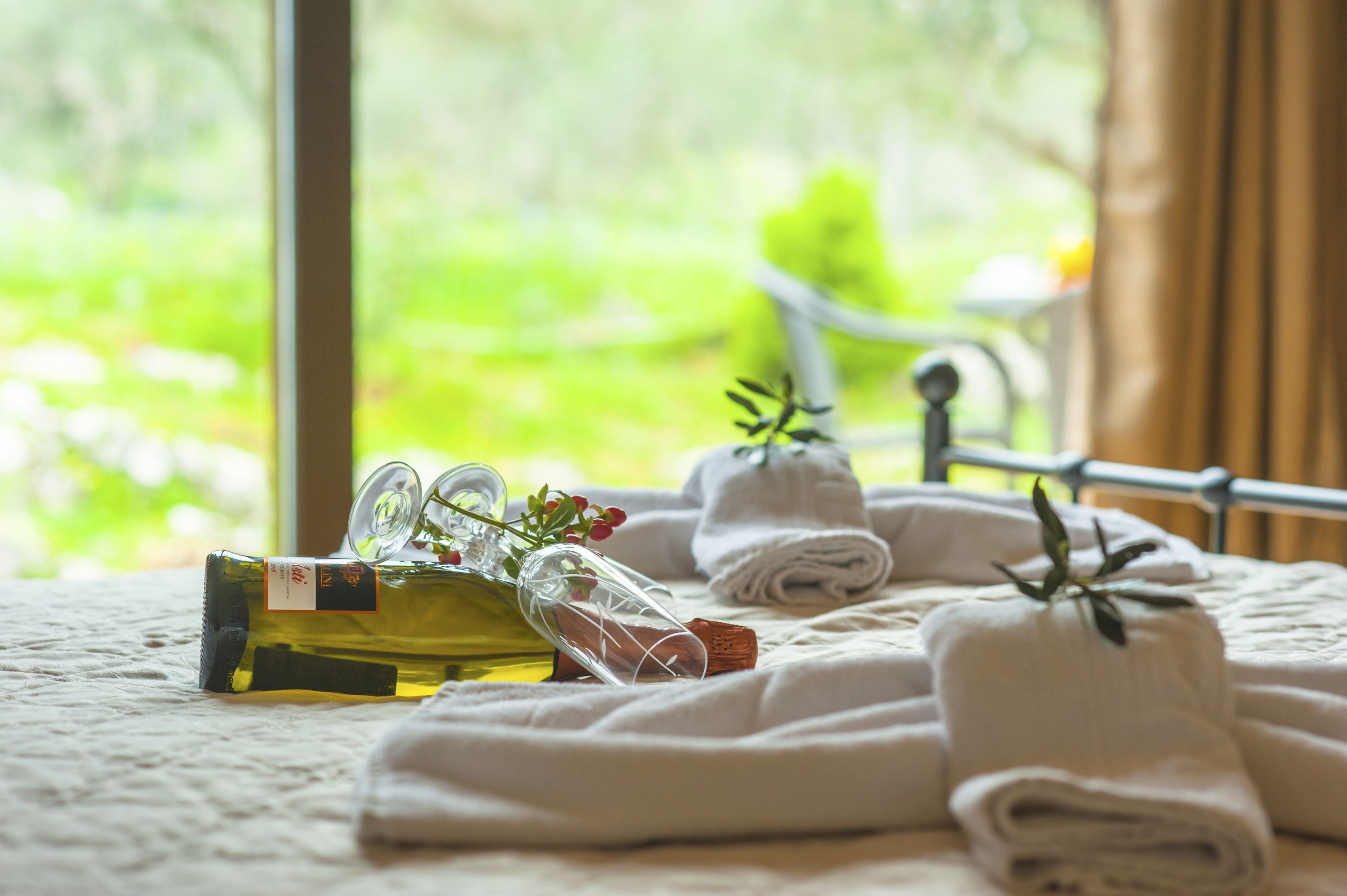 A neatly arranged bed with rolled towels, a bottle of olive oil, and a glass bottle with a cork, set against a window with a view of greenery.