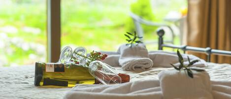 A neatly arranged bed with rolled towels, a bottle of olive oil, and a glass bottle with a cork, set against a window with a view of greenery.