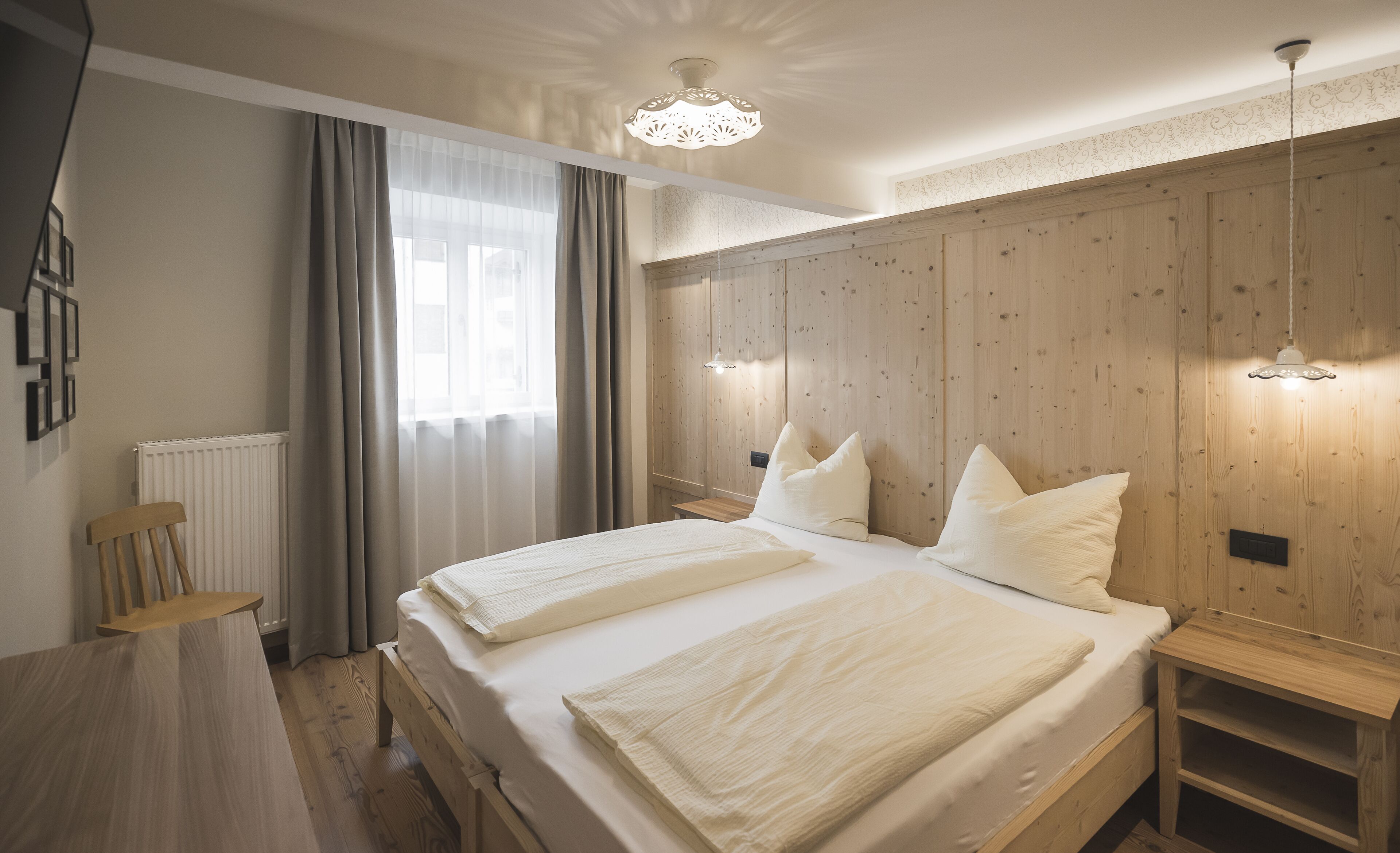 Un dormitorio con cabecera de madera, dos camas con ropa de cama blanca, una mesita de noche de madera, una silla de madera, un radiador y una ventana con cortinas de encaje.
