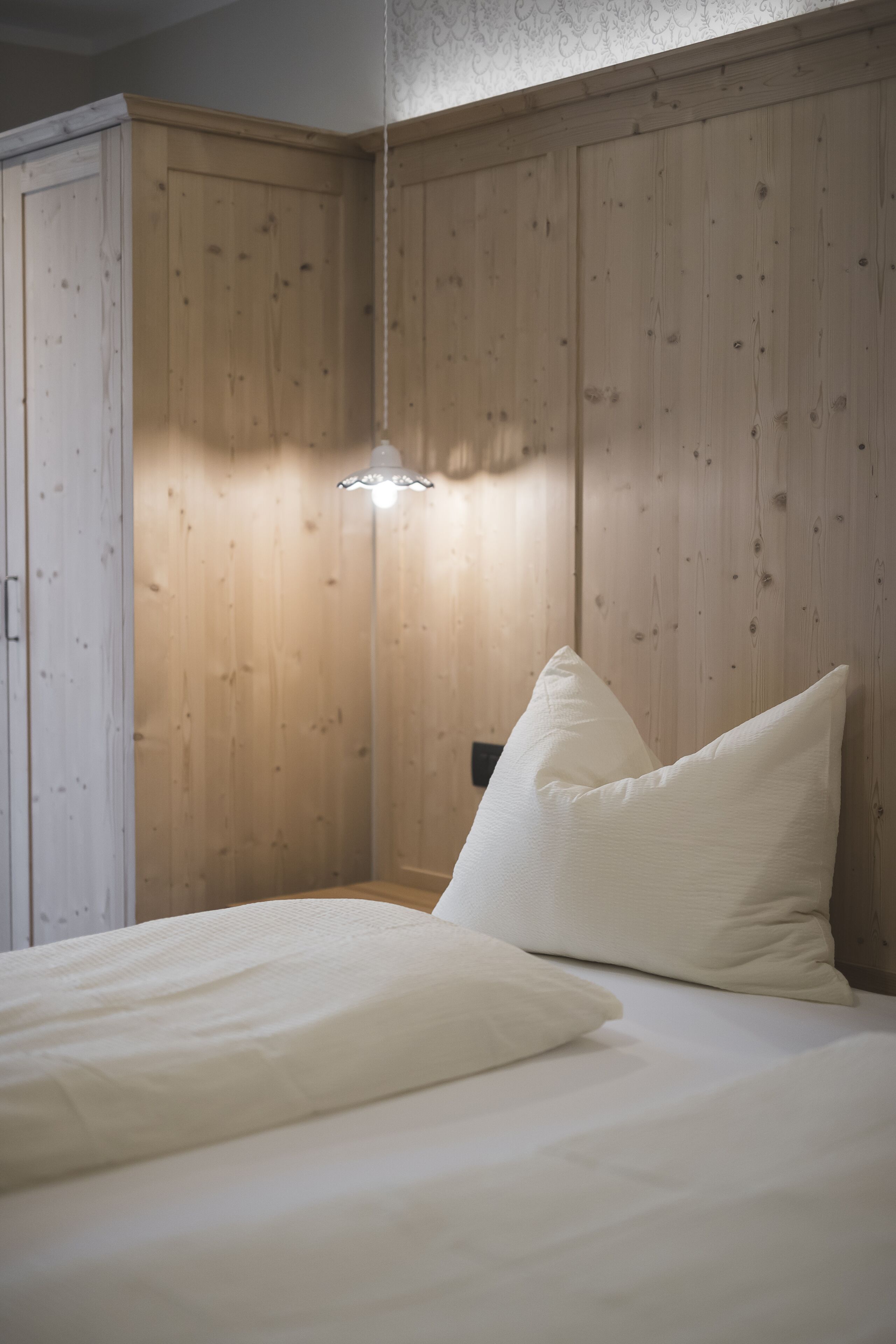 A bedroom with a wooden wardrobe, a bedside table, and a bed with white bedding.