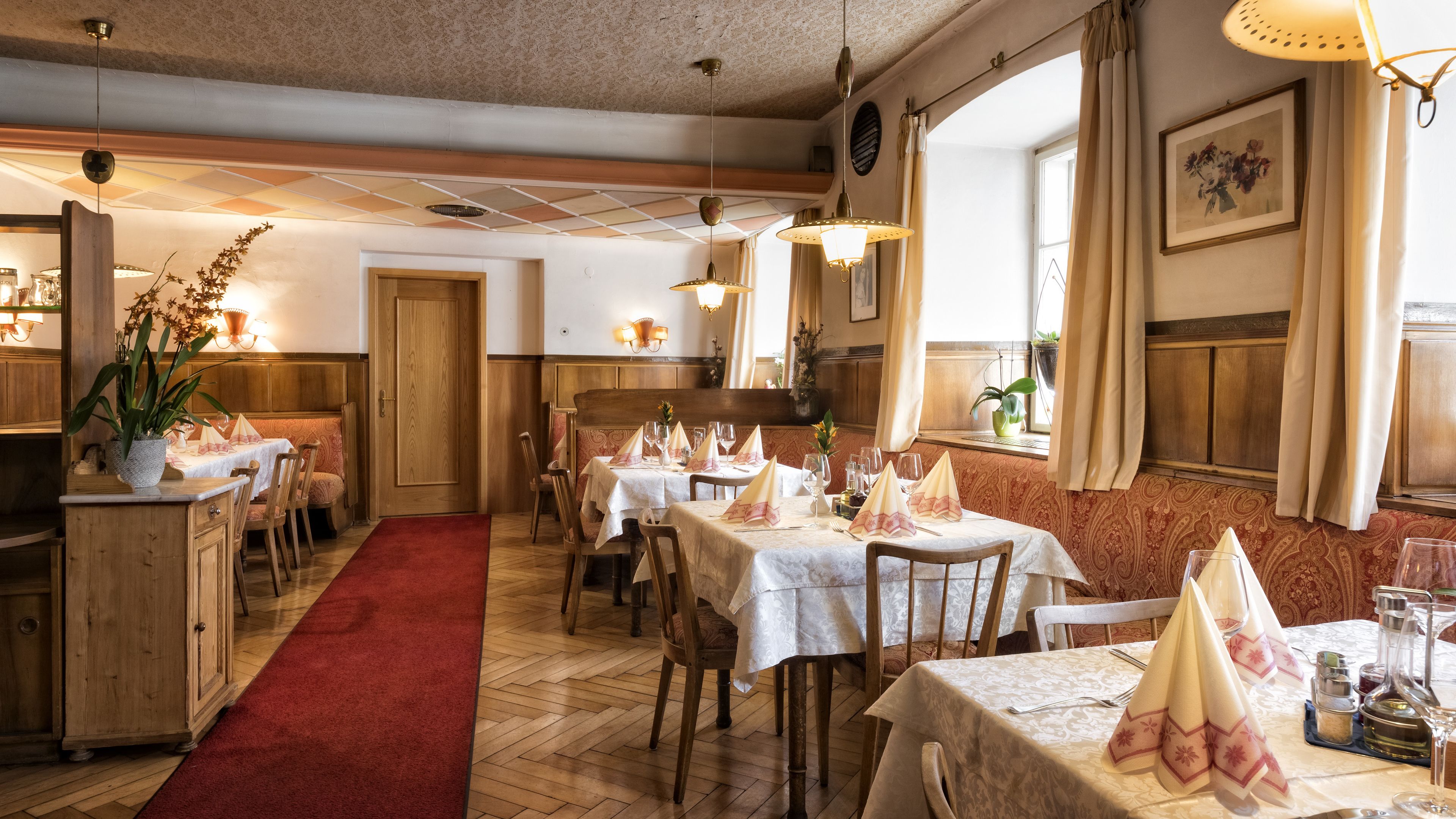 A restaurant with wooden furniture, white tablecloths, and a red carpet leading to the dining area.