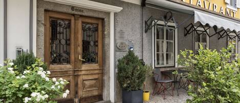 Entrada de hotel con puerta de madera, un letrero que indica un restaurante y mesas al aire libre con sillas.