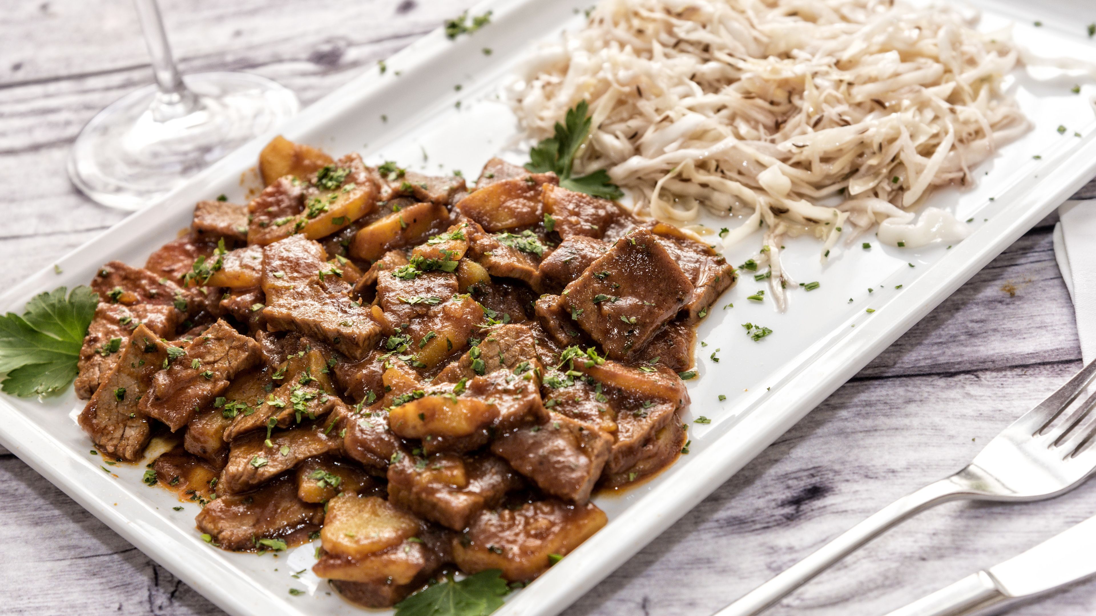 A rectangular white plate with a beef dish garnished with herbs, accompanied by a side of shredded white noodles and a glass of white wine on a wooden table.