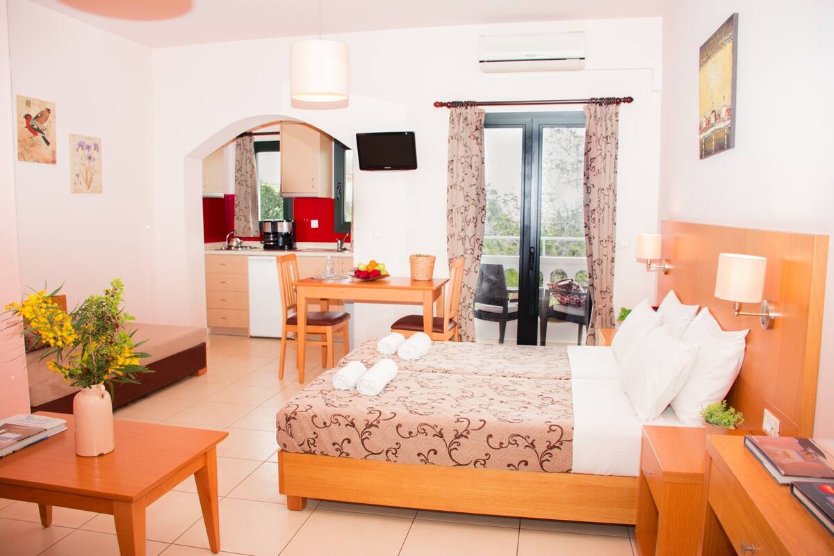 Una camera d'albergo con un letto, una scrivania, una sedia, un tavolo da pranzo e una zona cucina visibile tramite un arco.