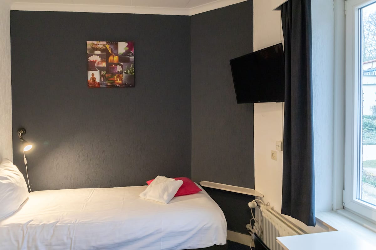 A hotel room with a bed, a wall-mounted TV, a window with curtains, and a radiator.