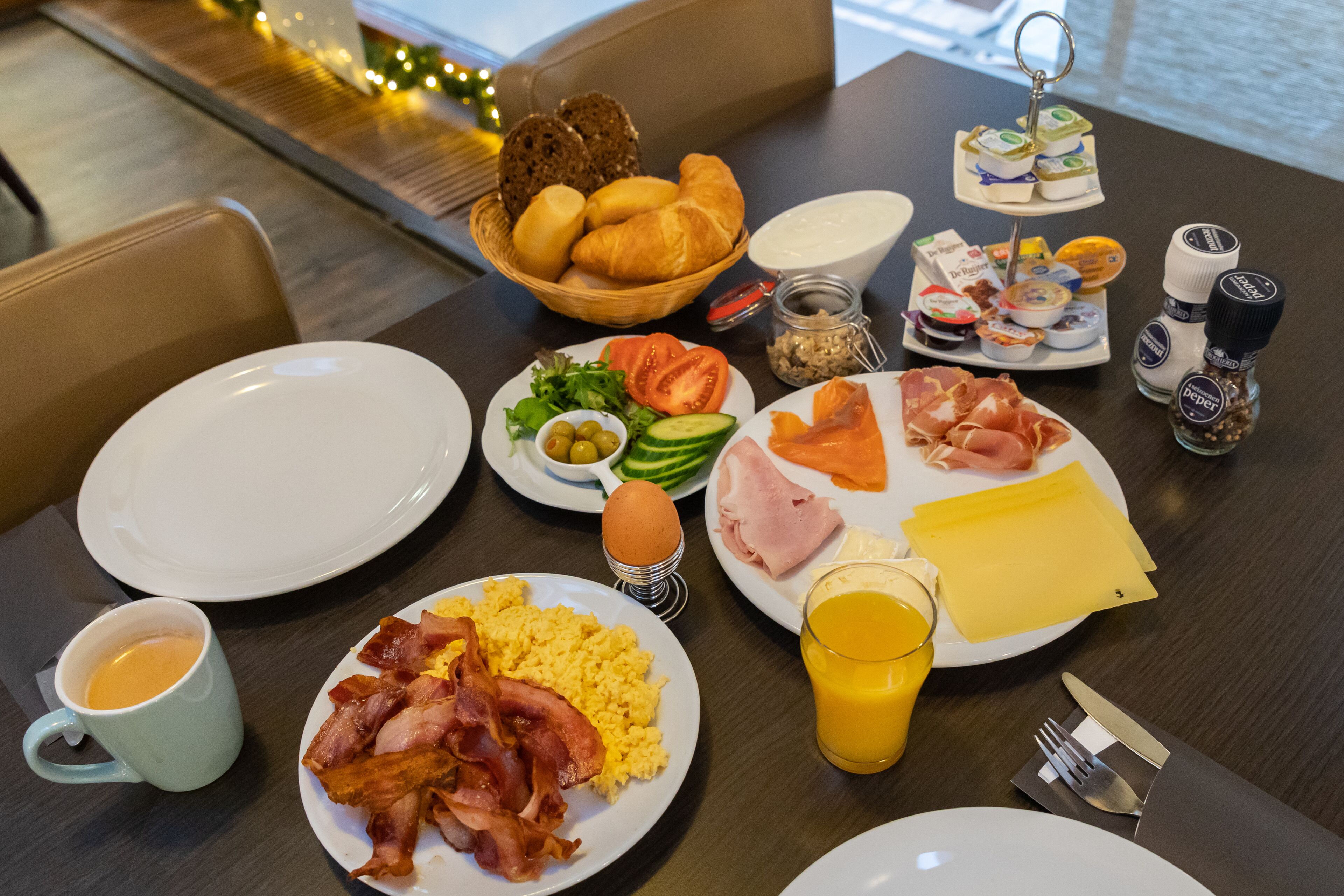 A breakfast table with plates of eggs, bacon, ham, cheese, croissants, and bread. There are also condiments, a glass of orange juice, and a cup of coffee.