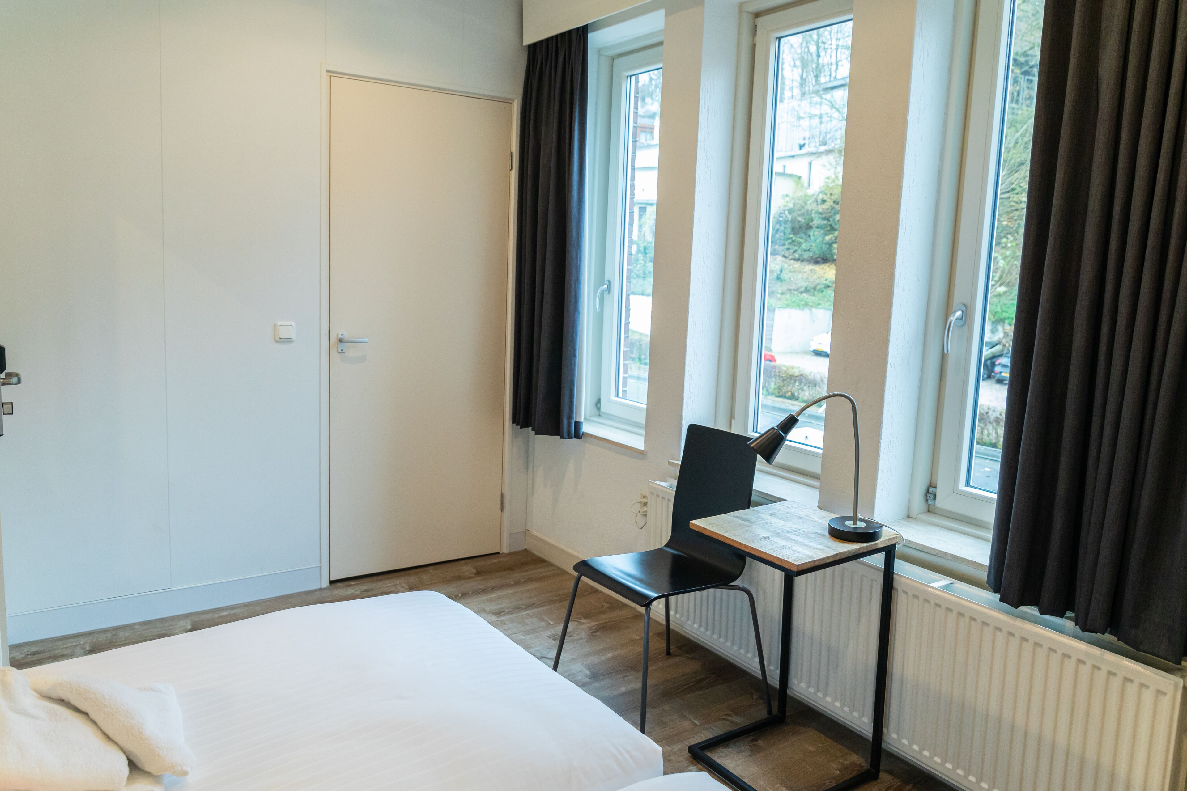 Een hotelkamer met een bed, een bureau met stoel, een raam met gordijnen en een radiator.