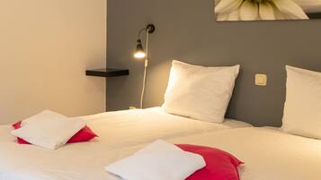 Een net opgemaakt bed met witte en rode kussens, een bedlampje en een wandplank.