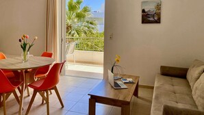 A living room with a sofa, a dining table with chairs, a laptop on a side table, and a view of palm trees through an open door. - Manolis Apartments (Agios Vasileios)