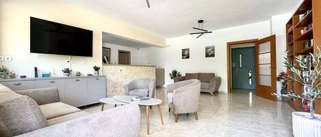 Una sala de estar moderna con un sofá gris, una mesa de centro y un televisor montado en la pared. Se observa una cocina al fondo, con un muro de ladrillo y una puerta de madera.