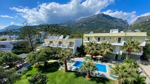 A resort with a swimming pool, palm trees, and mountainous views. - Manolis Apartments (Agios Vasileios)
