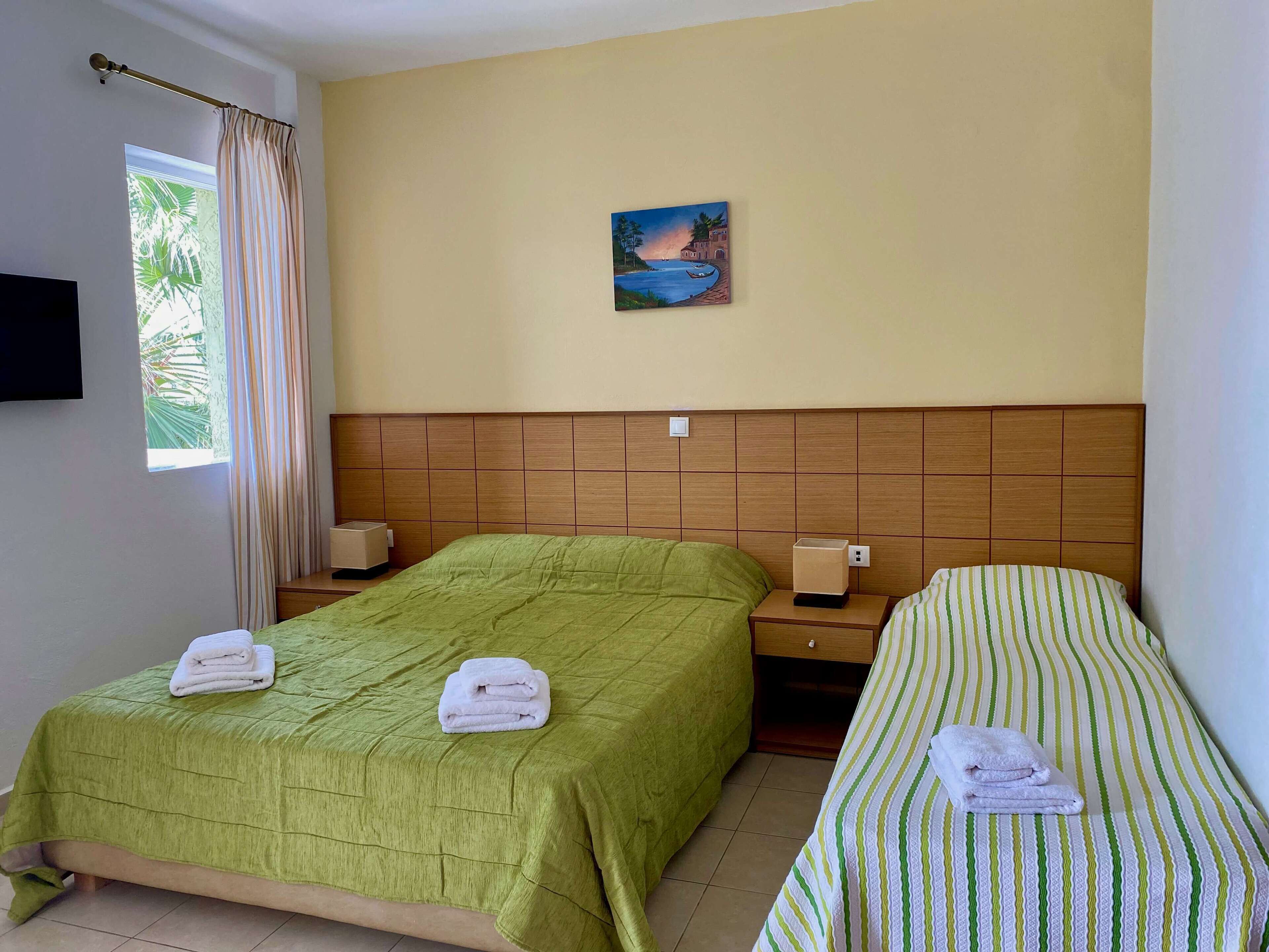 A hotel room with two single beds, each with a green and striped bedspread, and a painting of a coastal scene on the wall.