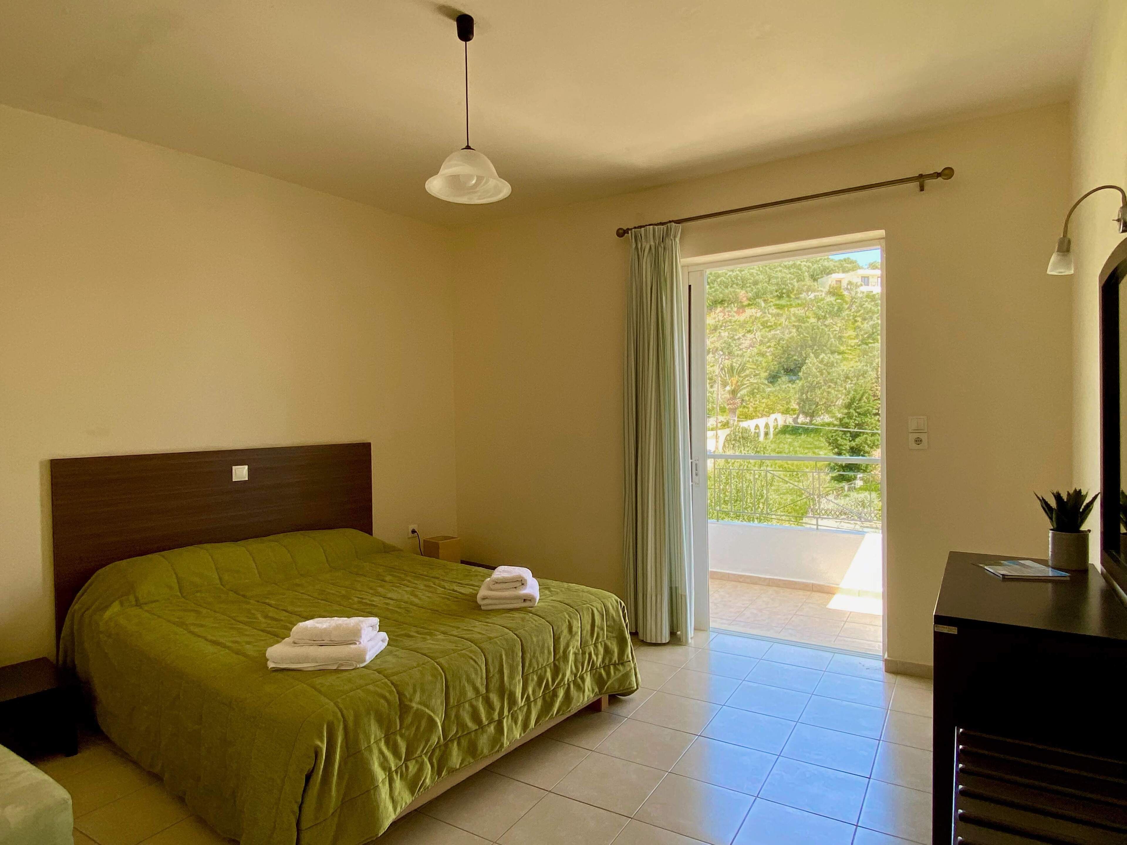 A hotel room with a bed, a desk, a chair, a window with a view of greenery, and a ceiling light.
