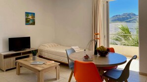 A modern room with a bed, a TV, a dining table with chairs, and a balcony with a view of the sea and mountains. - Manolis Apartments (Agios Vasileios)