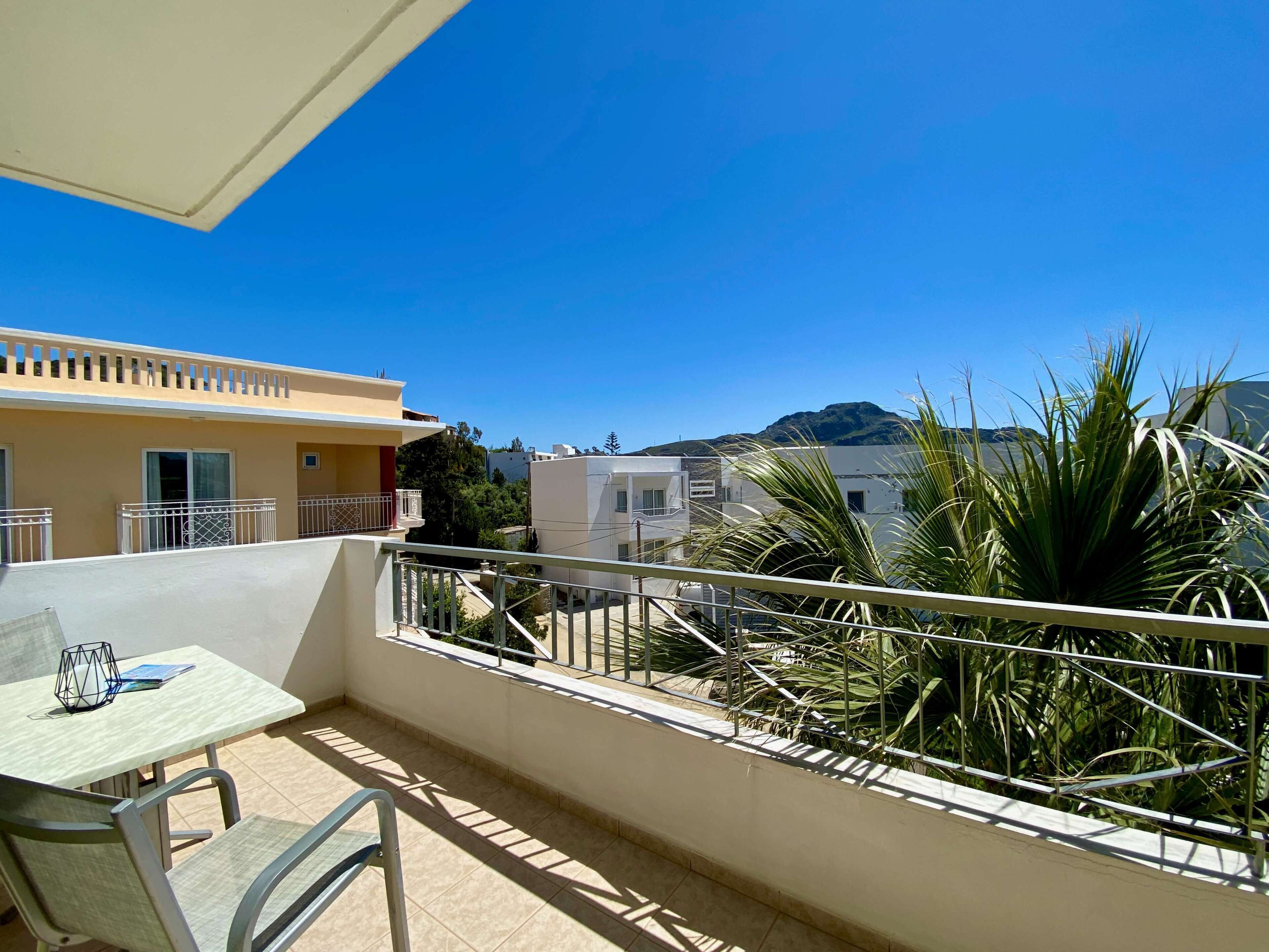 A balcony with a table and chairs, a railing, and a view of buildings and mountains in the background.
