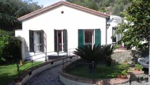 Un edificio blanco con contraventanas verdes, un camino de piedra que lleva a un banco de piedra circular y plantas en macetas.