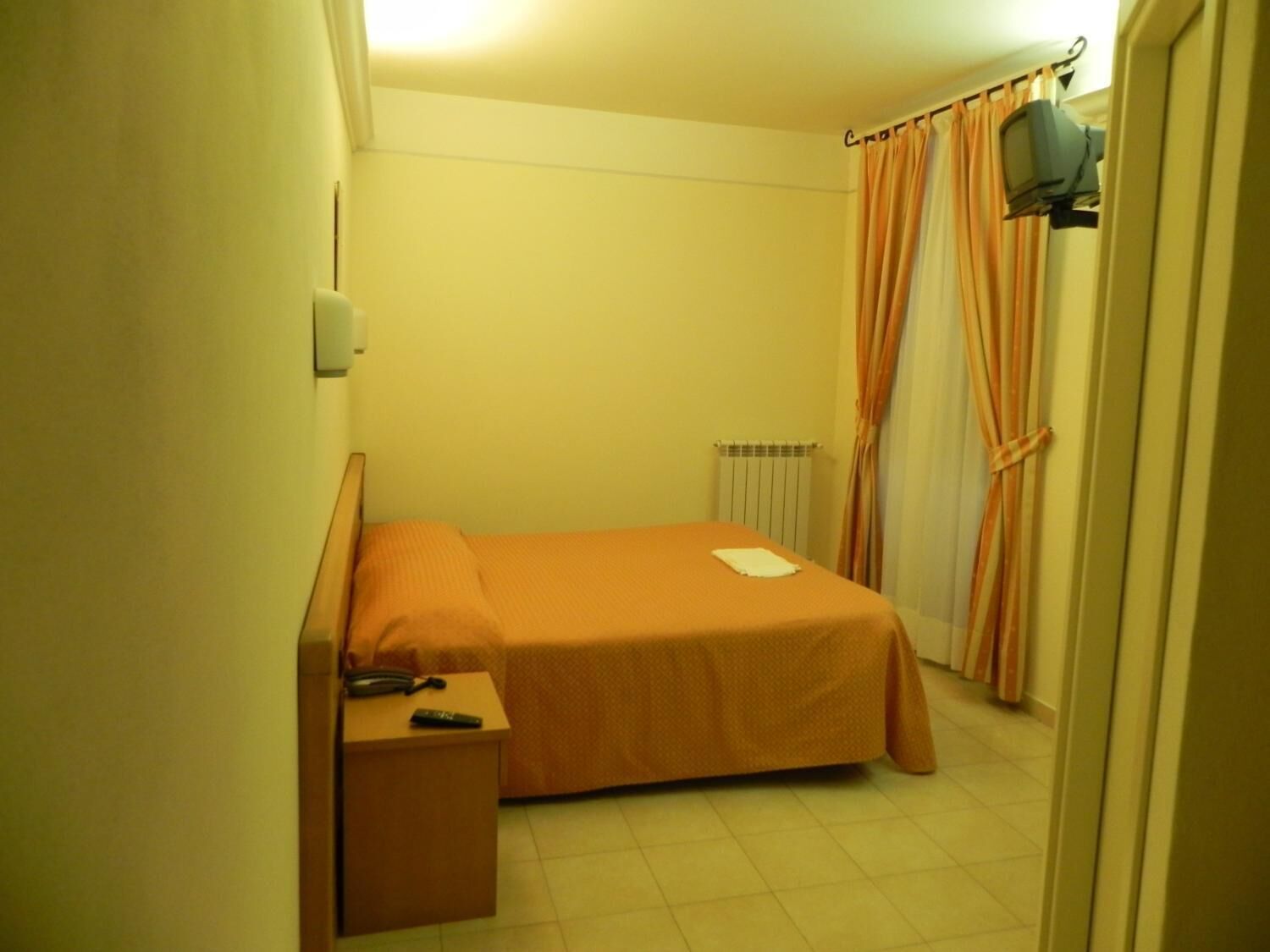 A small, single-bed room with a wooden headboard, a bedside table with a phone and remote control, a radiator, and curtains.