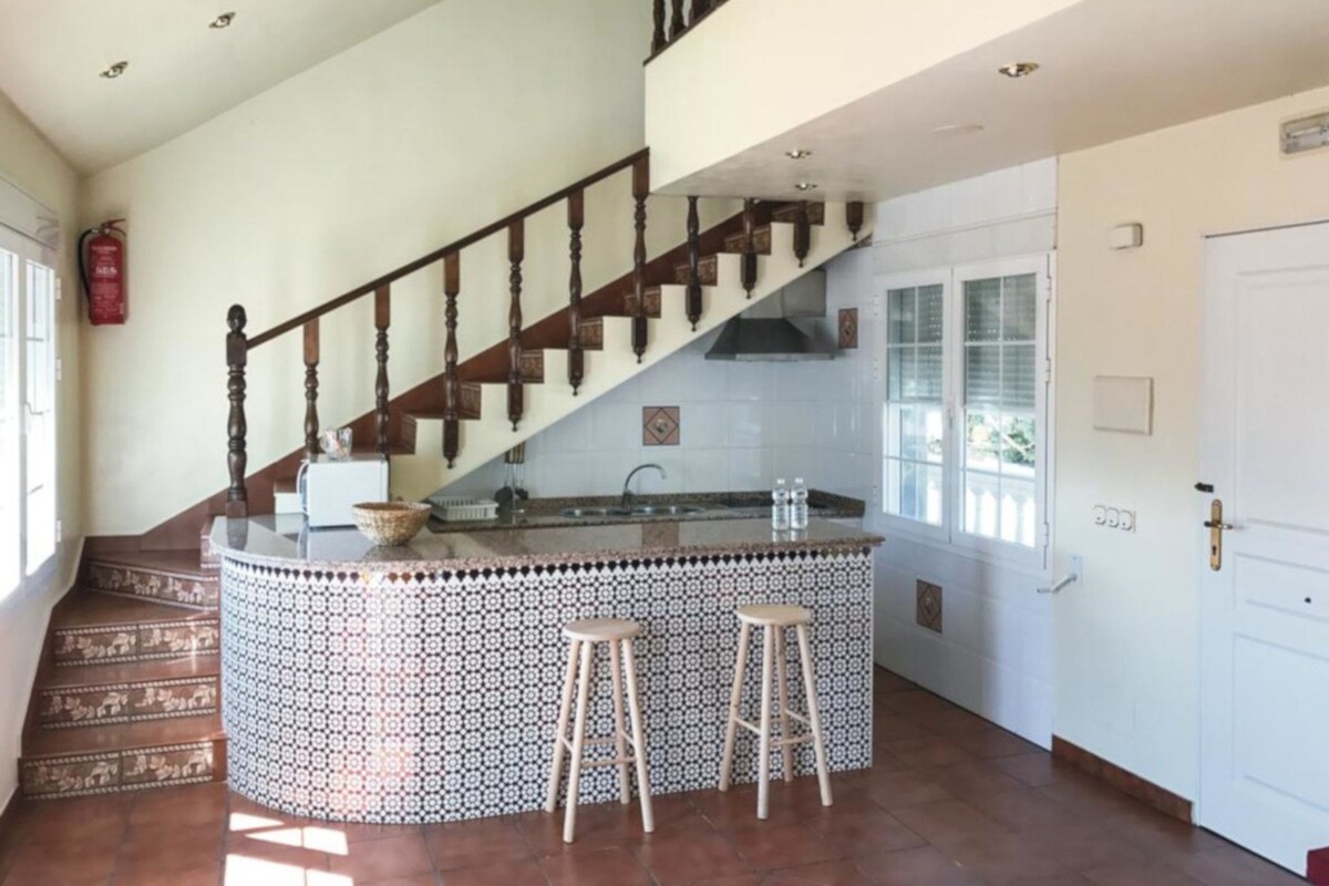 Une cuisine avec un îlot central, deux tabourets, un escalier et un extincteur.