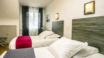 Habitación de hotel con dos camas, cabecero de madera, ventana con cortinas, luz de pared y una mesita con lámpara.