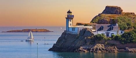 A coastal scene with a lighthouse, a sailboat, and buildings on a rocky cliff.
