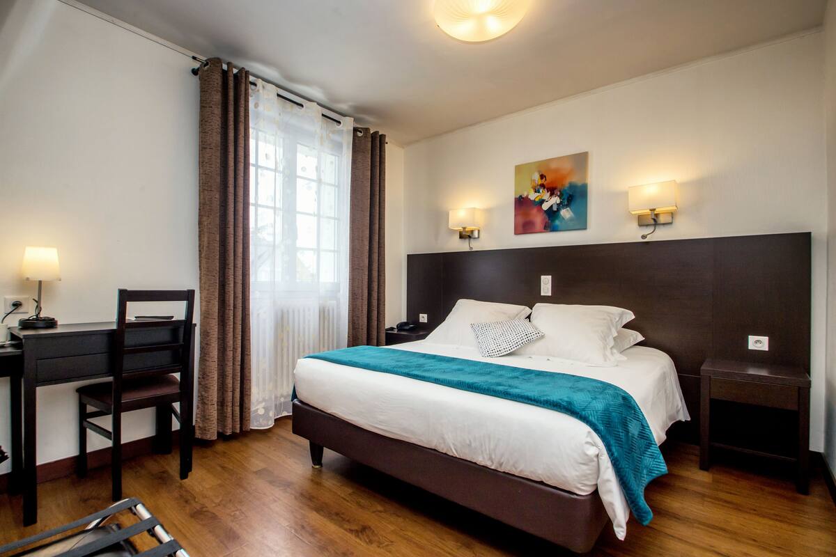 A hotel room with a bed, a desk, a chair, a nightstand, and a painting on the wall.