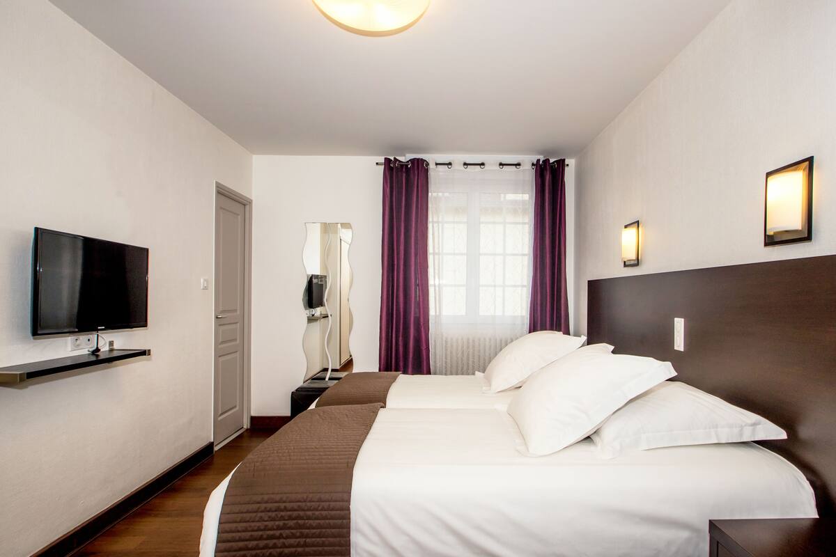 A hotel room with a TV on a shelf, a bed with white bedding, a wooden headboard, and a window with purple curtains.