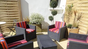 Ein Terrassenbereich im Freien mit Korbmöbeln, darunter Stühle und ein Tisch, umgeben von Topfpflanzen und einem Holzzaun.