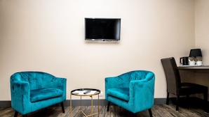 Ein Zimmer mit zwei blaugrünen Sesseln, einem kleinen runden Tisch, einem flachbildfernseher an der Wand und einem Schreibtisch mit Stuhl und Lampe.