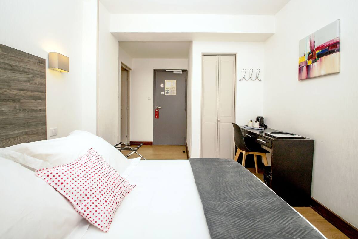 A hotel room with a bed, a desk, and a chair. There is a door leading to another room and a wall-mounted artwork.
